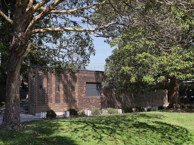 Exterior view of a low, rectilinear brick building partially screened by mature trees. The dark brown brick walls include a small horizontal louvred window, with lawn and landscaped planting in the foreground. The building has a restrained, institutional or modernist character and is photographed in bright daylight.