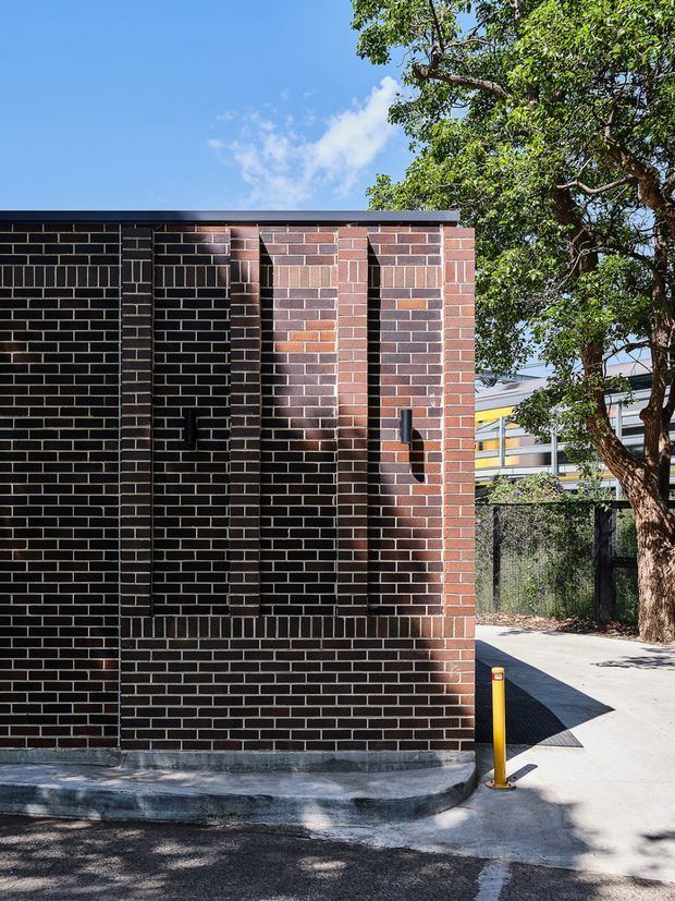 Exterior architectural photograph of a compact brick building or wall with deep vertical recessed bays and projecting brick fins. The dark and reddish brickwork creates a strong geometric facade, capped by a simple flat roof edge. A paved access path, yellow bollard, mature tree, and neighbouring structure are visible around the building.