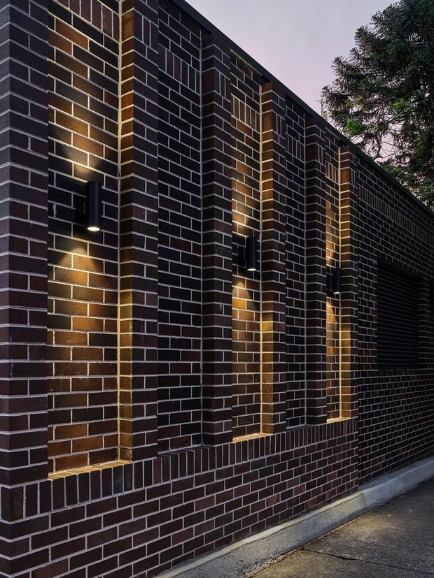 Close-up exterior view of a dark brown and charcoal brick facade with projecting vertical brick piers, recessed illuminated wall sections, black wall-mounted lights, and a dark garage or service opening. The image focuses on the building envelope and its textured masonry detailing in a moody dusk setting.