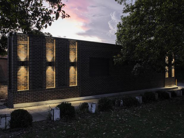 A low, contemporary dark-brick building viewed at dusk, with a long solid facade punctuated by narrow vertically oriented windows. Warm interior or concealed lighting glows through the window openings and washes the brickwork, while trees frame the building and a simple planted verge and paved walkway run along its base.