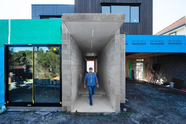 Photograph of a modern building under construction, showing a deep concrete-block entry passage or portal with a person walking through it. Dark vertical cladding and large glazed sliding doors are visible on the facade, while blue construction wrap, exposed ground, scaffolding materials, and an adjoining covered area indicate an active building site.