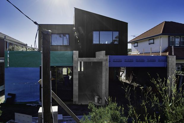 Exterior street view of a contemporary two-storey residential building under construction or renovation. The facade combines dark vertical cladding with exposed grey masonry/concrete elements, large rectangular windows, and a partially screened lower level. Construction materials and temporary fencing are visible in the foreground, with a neighbouring traditional weatherboard house beside it.