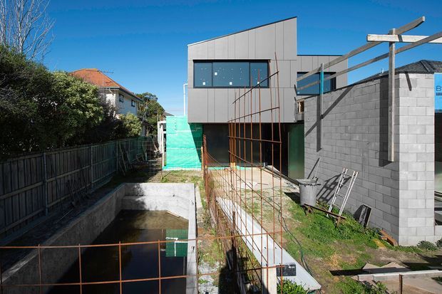 Exterior view of a contemporary multi-level building under construction, showing a box-like upper volume with grey panel cladding, large dark-framed glazing, exposed structural steel and timber framing, concrete retaining walls, and an unfinished lower level. Adjacent residential properties and fencing are visible around the narrow sloped site.