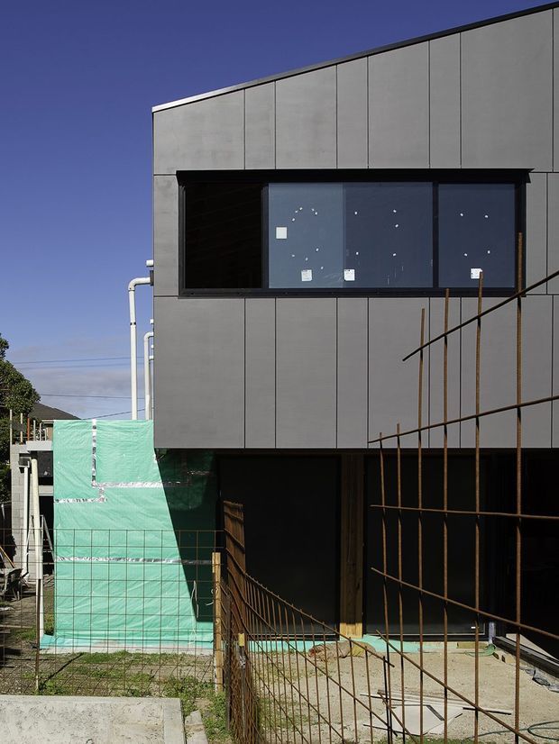 Exterior photograph of a contemporary multi-level building under construction. The facade is clad in large dark grey rectangular panels with narrow vertical joints, a projecting black-framed glazed window band, and a deep recessed lower-level opening. Temporary green construction sheeting, exposed reinforcing steel, scaffolding elements, and unfinished concrete paths indicate an active construction site. Clear blue sky and neighbouring structures are visible in the background.