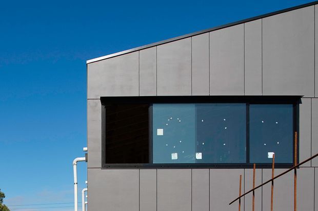 Exterior view of a contemporary box-shaped building clad in large grey panels. A projecting black-framed glazed window bay interrupts the facade, with visible temporary labels on the glass. The image shows a clean, minimal facade against a clear blue sky; exposed service pipes and a simple metal stair or railing are visible at the edges.