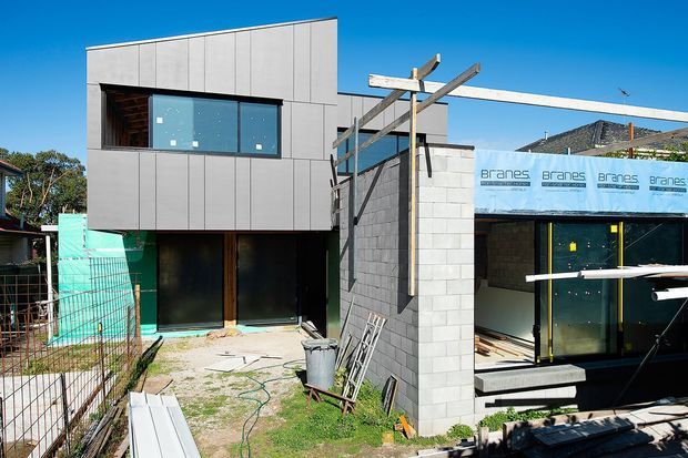 Exterior view of a contemporary multi-level residential building under construction. The facade combines dark grey vertical panel cladding with exposed grey concrete blockwork, large glazed windows and doors, and a partially framed upper-level structure. Construction materials, scaffolding, temporary fencing, and unfinished landscaping are visible around the site beneath a clear blue sky.