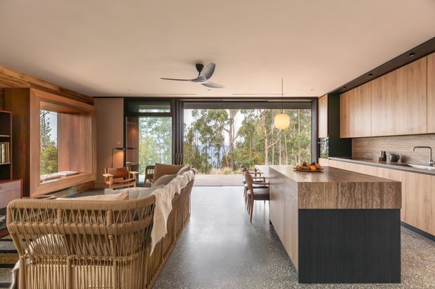 Open-plan contemporary kitchen and living area with extensive timber cabinetry and wall panelling, a large stone island, polished concrete or terrazzo-style flooring, dark-framed floor-to-ceiling glazing, woven lounge seating, and a view into a wooded landscape. A ceiling fan and pendant light add functional and decorative elements to the warm, refined interior.