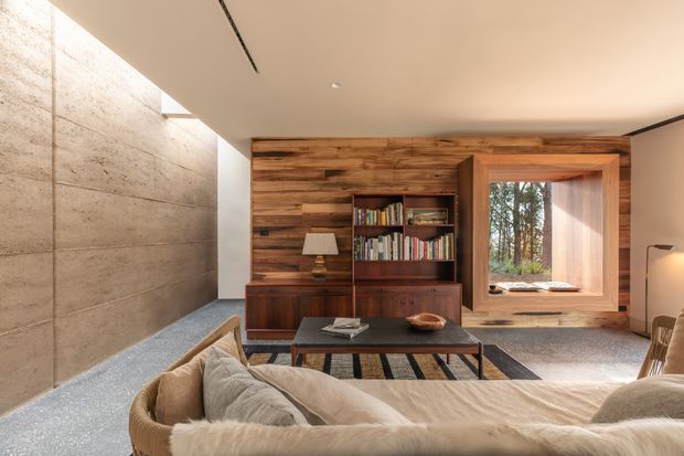 Warm, refined living room with a low-profile lounge arrangement facing a built-in timber feature wall. The wall incorporates dark timber cabinetry and an open bookshelf, while a large timber-framed picture window looks onto trees. A concrete or stone-textured wall runs along the left, with a polished dark coffee table, soft neutral seating, and a partially visible rug. The ceiling is minimal and includes a narrow linear slot detail.