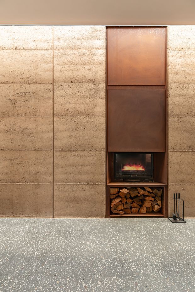 Close-up interior view of a built-in fireplace wall. Large textured beige stone or concrete panels frame a recessed wood-burning fireplace with a dark metal surround, copper-toned metal cupboard panels above, and an open log-storage niche below. A terrazzo-style floor extends across the foreground, with fireplace tools placed to the right.