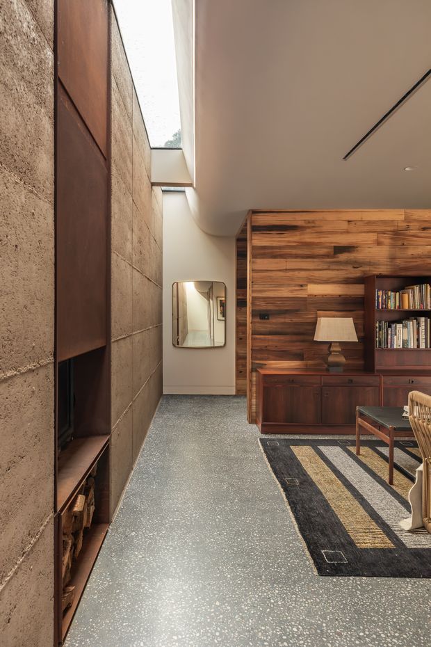 A refined, warm-toned living room or lounge viewed along a circulation area. The space features a textured concrete-block wall with a dark built-in fireplace and timber storage, a large glazed skylight or clerestory opening, a richly finished reclaimed-looking timber feature wall, and dark timber built-in shelving and cabinetry. A mid-century style lounge chair, table lamp, books, and geometric black-and-neutral rug complete the interior. The polished terrazzo-like floor and restrained linear ceiling lighting create a modern, gallery-like atmosphere.