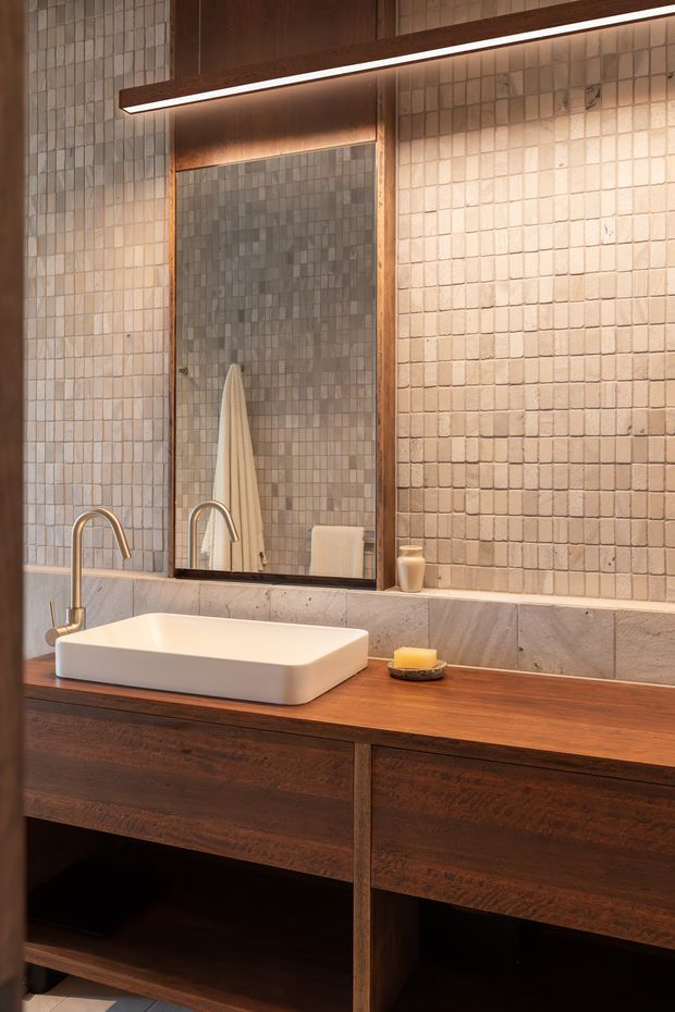 A warm, contemporary bathroom vanity featuring a white vessel sink with a brushed metal tap set into a dark timber benchtop and custom timber cabinetry. The wall is finished with small square textured beige and grey tiles, with a large timber-framed mirror and a slim linear wall light above it. Neutral stone surrounds and minimal accessories create a refined, earthy atmosphere.