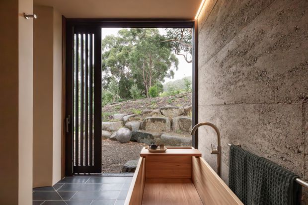 A compact, warmly lit kitchen or utility-style space with a timber-lined trough sink and tall curved faucet in the foreground. The room features dark tiled flooring, a dark-framed glazed door with vertical screening, a rough textured concrete or stone wall, integrated linear ceiling lighting, and a heated towel rail. Through the open doorway is a landscaped rocky garden with mature trees.