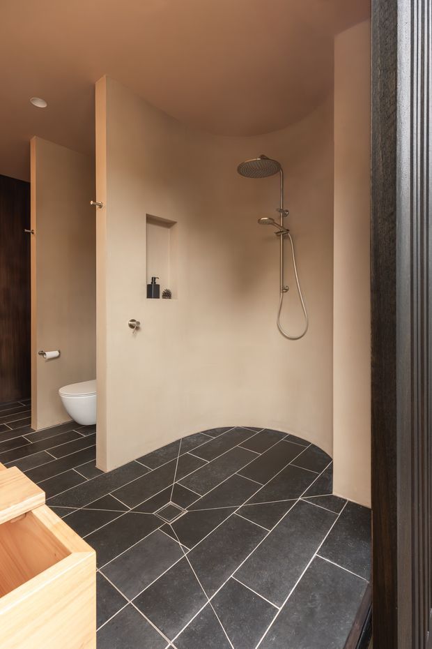 A warm, sculptural bathroom with a curved plaster shower enclosure, exposed rainfall shower and handheld shower, recessed wall niche, wall-mounted controls, and a partially visible toilet. The floor is finished in large dark charcoal tiles with contrasting light grout arranged in a geometric pattern. Timber cabinetry or a bath surround is visible in the foreground, and a dark vertical timber-toned screen frames the right side.