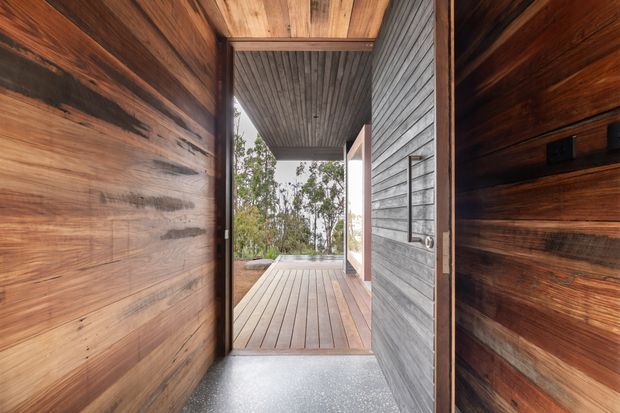 View through a timber-lined entry passage toward a covered outdoor deck. Dark and warm-toned wood wall and ceiling linings frame the opening, while a grey horizontal-clad wall and timber decking lead toward a wooded landscape. The image emphasizes a sheltered indoor–outdoor threshold and natural material palette.