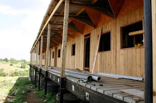 A long raised timber building under construction, with exposed timber wall cladding, open window and door openings, a covered verandah supported by timber posts, and visible roof framing. Construction materials and pipes are stored along the unfinished deck. The structure sits elevated above the ground in a rural-looking setting.