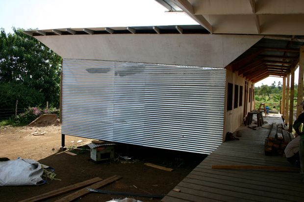 Exterior view of a partially constructed or rustic building with a large corrugated metal-clad wall, deep roof overhangs, exposed timber roof framing, and a raised timber walkway or deck along the side. The setting appears rural and tropical, with construction materials and unfinished ground visible.