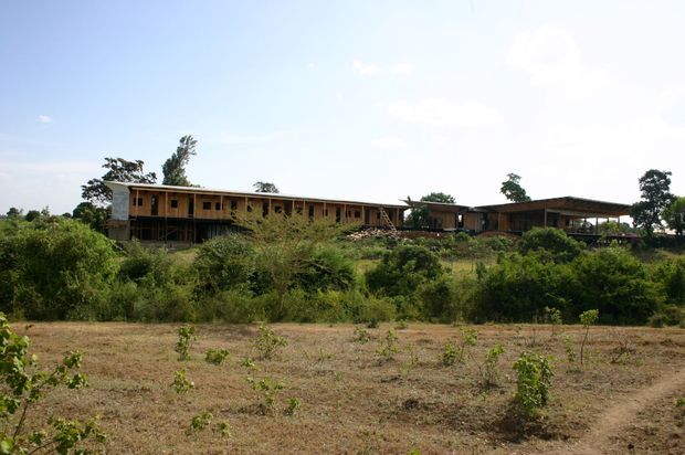 Wide exterior view of a long, low-profile building set within a dry, lightly cultivated landscape and dense surrounding vegetation. The structure has a simple horizontal form, exposed timber-toned wall surfaces, a broad low-pitched roof, and an open-sided covered area at one end. The image is photographed from a distance under a bright sky.