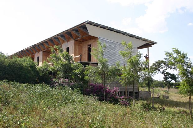 Exterior view of a raised, modern tropical-style house set within a landscaped rural garden. The building features extensive timber elements, a broad overhanging pitched roof with exposed rafters, a projecting balcony, and a corrugated metal-clad side wall. Dense planting partially screens the lower level.