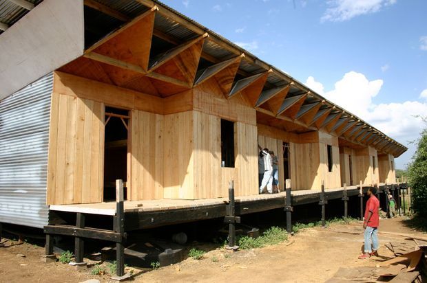 A long, raised single-storey building under construction, clad primarily in vertical timber boards with sections of corrugated metal siding. The structure sits on elevated supports above bare ground and features a prominent repetitive exposed timber roof structure with corrugated roofing. Openings for doors and windows are visible, along with several people on the unfinished platform and beside the building.