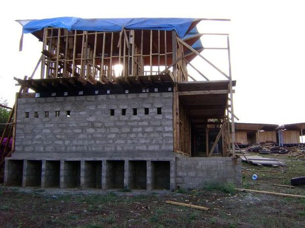 Partially constructed two-storey building on an unfinished site, with exposed concrete-block lower walls, timber framing and scaffolding, and a blue temporary tarp covering the upper structure.
