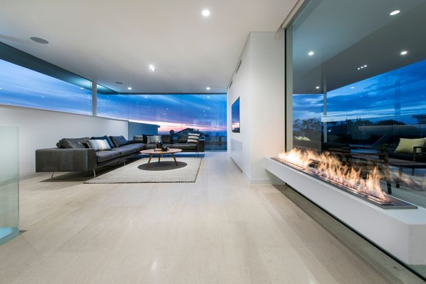 A spacious contemporary living room at dusk, featuring a large dark sectional sofa, round coffee table, pale flooring, and expansive floor-to-ceiling glazing with panoramic views. A long linear gas fireplace is integrated into a low white built-in wall beneath the right-hand glazing. Recessed ceiling lights and a clean, minimal interior create a refined modern atmosphere.