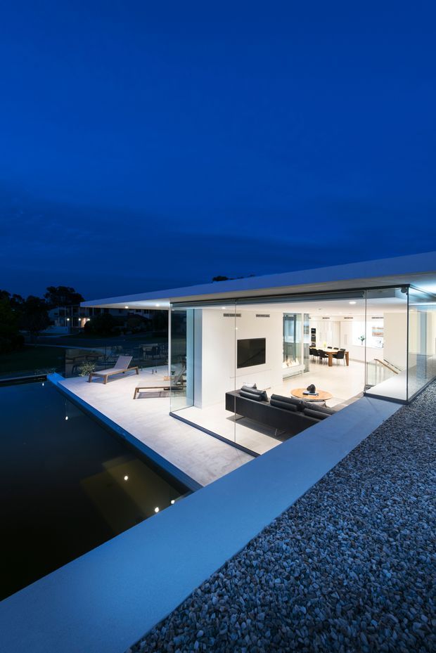 Dusk exterior view of a sleek, single-storey modern residence arranged around a dark reflecting pool. Extensive floor-to-ceiling glass reveals a bright open-plan living room and dining/kitchen area, while a pale paved terrace with outdoor loungers projects over the water. The flat roof is finished with a gravel-covered roof surface, and strong blue evening light creates a dramatic contrast with the illuminated interior.
