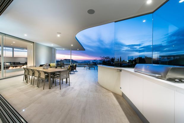 A spacious contemporary open-plan kitchen and dining area with full-height curved glazing overlooking a waterfront or coastal landscape at dusk. The room features a long dining table, streamlined white built-in cabinetry, an integrated stainless-steel barbecue or cooking appliance, pale stone-look flooring, recessed lighting, and a sculptural curved ceiling detail. The glazing creates a strong indoor-outdoor connection and panoramic sunset views.