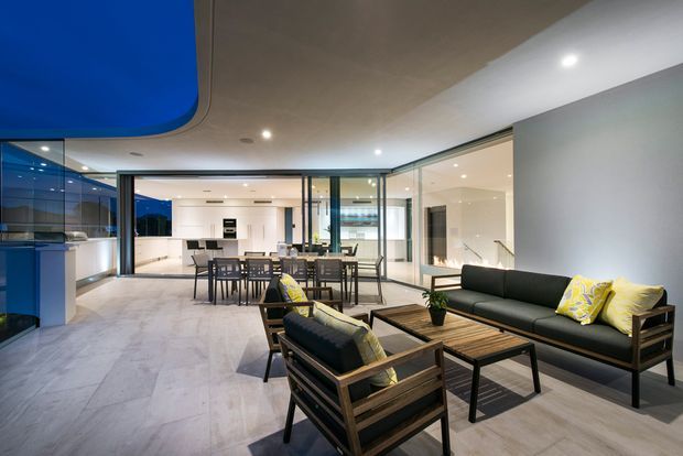 Open-plan contemporary interior combining a living area, dining area, and kitchen. The room features polished light stone or porcelain flooring, extensive black-framed glass partitions, white cabinetry and walls, timber-framed lounge seating, and a distinctive curved blue ceiling feature. The space is bright, spacious, and visually connected through transparent glazing.