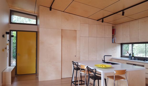 A compact, warmly lit contemporary kitchen with extensive pale timber cabinetry and matching timber wall and ceiling panels. The room includes a white island with integrated sink and black tap, a rear benchtop beneath a wide window, open shelving, track lighting, and a small dining or breakfast area with stools. A mustard-yellow entry door adds a strong accent colour.