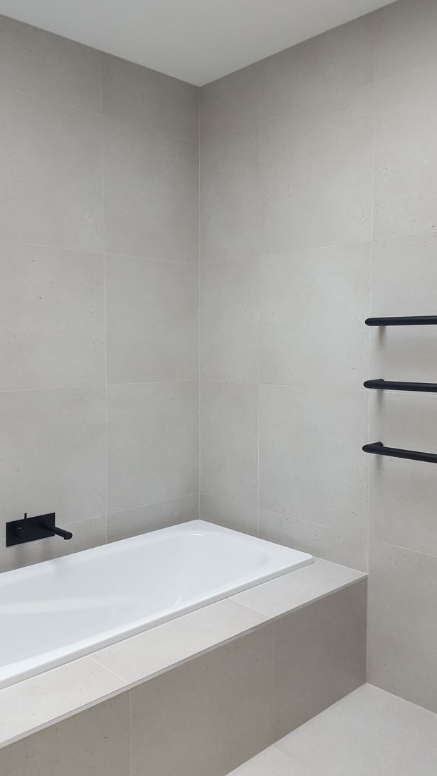 Minimal contemporary bathroom featuring a white built-in bathtub set into a pale stone or porcelain-tiled surround. Large light grey-beige wall and floor tiles create a restrained finish, with matte black wall-mounted tapware and three black heated towel rails providing contrast.