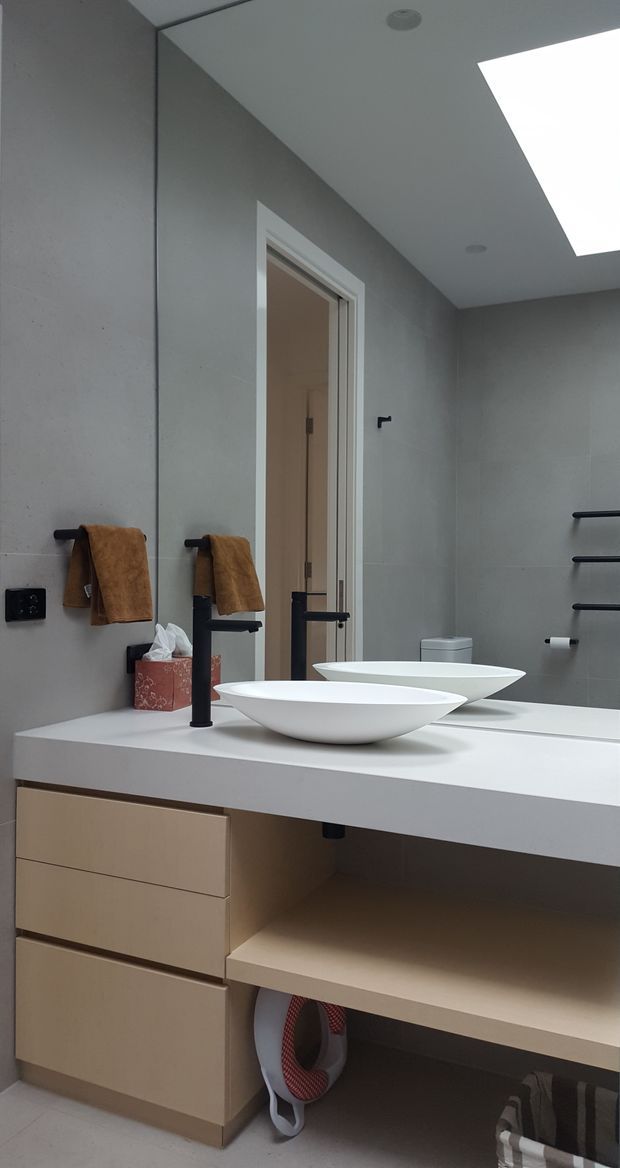 Contemporary bathroom vanity with a wide white benchtop, two sculptural white vessel basins, matte black tapware and hardware, and light timber-toned cabinetry. Large grey wall tiles, a full-height mirror, towel rails, open shelving, and a skylight create a clean, softly lit interior.