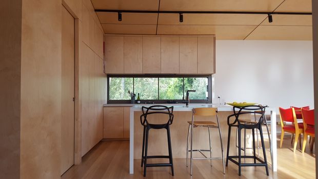 A warm, open-plan kitchen and dining area with pale timber cabinetry, a long island bench, bar stools, and a dining table with colourful red and yellow chairs. A horizontal window above the kitchen counter brings in greenery and natural light. The ceiling has timber panels with exposed black track lighting, and the room features timber flooring and built-in timber wall storage.
