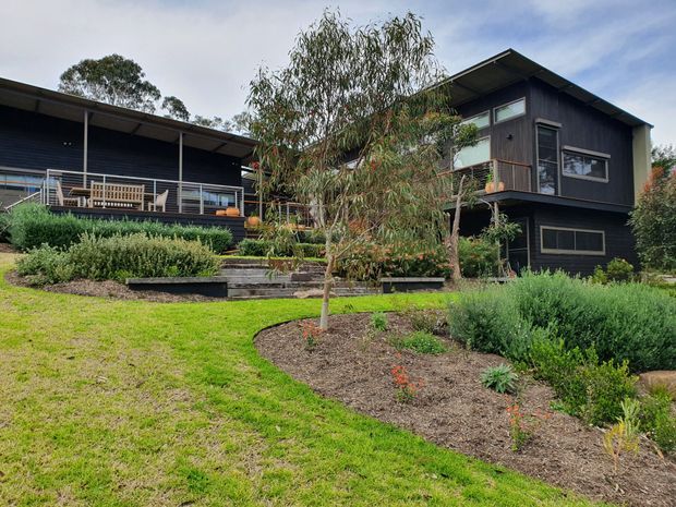 Exterior view of a modern, dark-clad multi-level house set within a landscaped garden. The home features broad roof overhangs, a raised upper volume, large windows, a covered deck with outdoor seating, horizontal cladding, and planted garden beds on a gently sloping site.