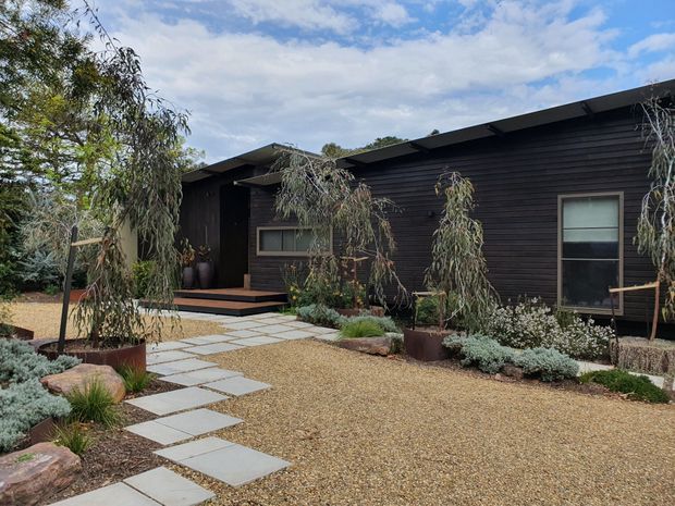 Single-storey dark-clad modern home viewed from a landscaped front courtyard. The low-pitched roof has broad overhangs, and horizontal dark timber-like cladding covers the facade. A pale stepping-stone path crosses a gravel forecourt with sculptural drought-tolerant planting, rocks, and slender trees. Simple rectangular windows and a recessed entry reinforce the restrained contemporary design.