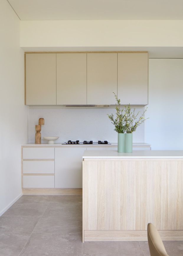 A bright, minimalist contemporary kitchen with pale timber-grain island cabinetry, white integrated wall cabinets, a light benchtop, gas cooktop, and a subtle pale terrazzo or stone backsplash. Large-format grey-beige floor tiles and soft white walls create a calm, restrained palette. A muted green vase with delicate branches provides the main accent.