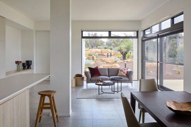 Open-plan contemporary interior combining a minimalist kitchen, dining area, and living room. The space features pale cabinetry, large dark-framed windows, light tiled flooring, a grey sofa, a dark dining table, and strong indoor-outdoor views toward a landscaped garden.