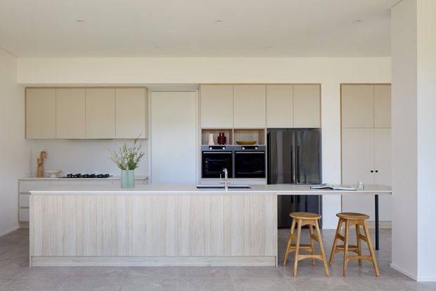 Bright contemporary kitchen with a long central island, pale timber-look vertical cabinetry, light neutral overhead cupboards, integrated black ovens, and stainless-steel refrigerator. Two timber stools sit at the island overhang, with subtle decorative greenery and a gas cooktop along the rear bench. The room has a clean, open, minimal composition and a light tiled floor.