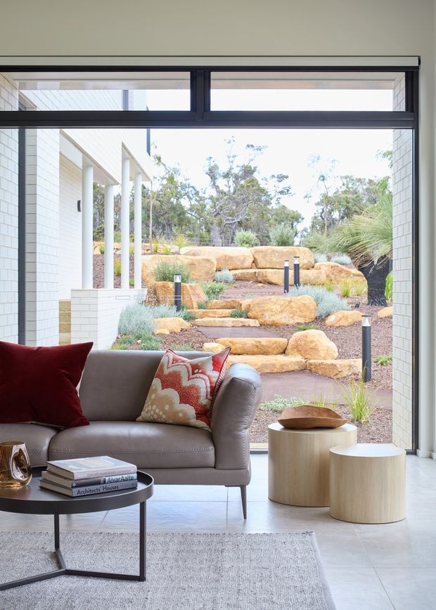 Bright contemporary living room opening directly onto a landscaped garden courtyard through large black-framed glazing. The room features a grey leather sofa with red and patterned cushions, a black round coffee table, pale cylindrical timber side tables, a light rug, and pale tiled flooring. White brickwork and slender columns are visible outside, with stepped sandstone landscaping and integrated pathway lighting.