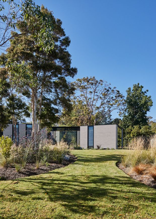 A low, single-storey contemporary pavilion-style house set within a landscaped garden. The building has dark grey horizontal cladding or masonry walls, extensive floor-to-ceiling glazing, slim dark window frames, and a flat roof. A broad lawn path leads through ornamental grasses and established trees toward the house under clear blue sky.