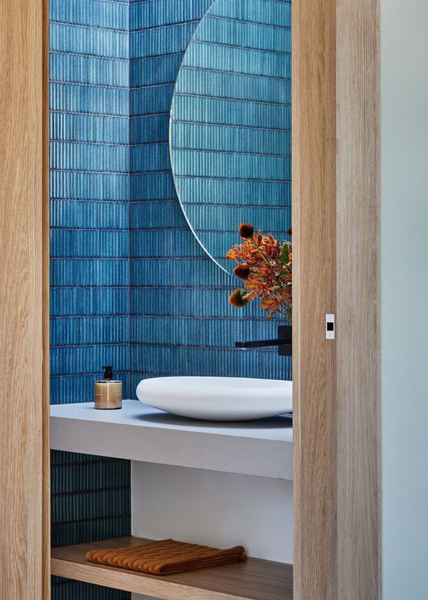 A contemporary bathroom vanity viewed through a timber-framed opening. The space features striking vertically ribbed blue wall tiles, a pale stone benchtop, a white vessel basin, a black wall-mounted tap, open timber shelving, and a large rounded mirror reflecting the blue tiled wall. A small sculptural floral arrangement and soap dispenser add restrained decorative accents.