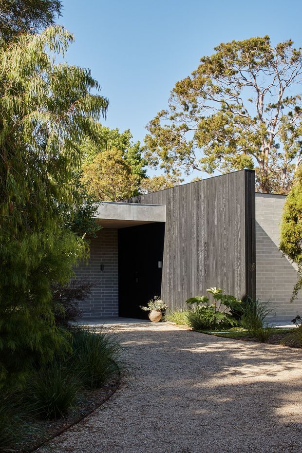 Exterior view of a low, contemporary house entrance set within landscaped gardens. The facade combines grey brickwork with a tall vertically textured dark timber-clad feature wall, a deep recessed black entry door, and simple flat roof forms. A pale gravel pathway leads through native and ornamental planting beneath mature trees in bright daylight.