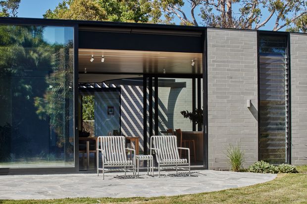 Single-storey modern residential facade opening onto a paved outdoor terrace. The composition features dark-framed expansive glazing, a recessed glazed entry or living area, a light grey painted brick wall, a tall narrow louvred window, and a warm timber-lined soffit. Two outdoor lounge chairs sit on the terrace, with established trees and low planting around the building.