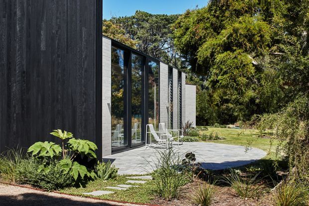 A low, modern pavilion-like building sits within a landscaped garden. The facade combines dark vertical timber cladding, pale masonry or concrete-block walls, and tall black-framed glazed doors or windows. A broad pale paved terrace with outdoor chairs fronts the building, while mature trees and native-style planting surround the structure.