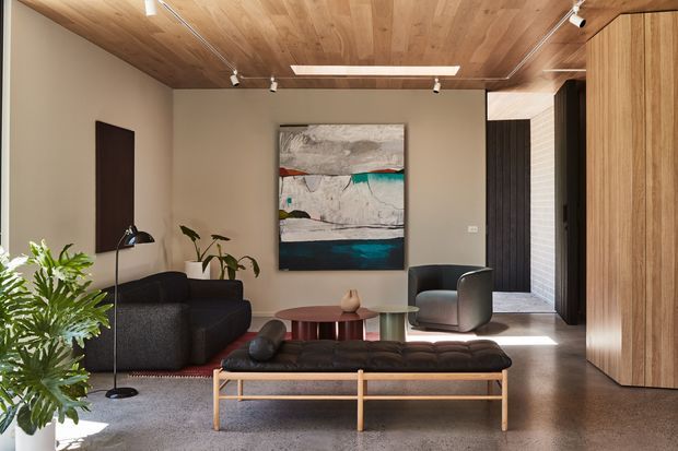A refined contemporary living room with a low black sectional sofa, upholstered lounge chair, timber-framed coffee table, abstract artwork, indoor plants, and a sculptural floor lamp. The room features warm timber wall and ceiling lining, a dark polished floor, neutral plastered walls, recessed and track lighting, and a timber-lined opening leading to a hallway or entry area.