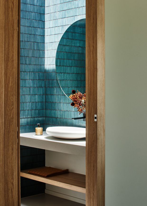 A refined bathroom vanity viewed through a timber-framed doorway. The compact space features deep teal vertically ribbed wall tiles, a white stone countertop, a sculptural white vessel basin, open timber shelving, a round mirror, and a small wall-mounted tap. The muted green adjacent wall and warm timber framing create a moody, contemporary atmosphere.