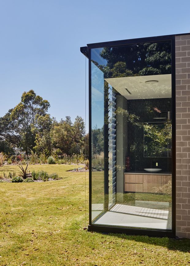 Exterior view of a contemporary house extension with a tall, black-framed glazed corner enclosing a bathroom. The bathroom includes a floating timber vanity, dark textured wall finish, pale ceiling, and a rounded basin, with strong visual connections to the landscaped garden. A dark brick wall forms the right edge of the facade.