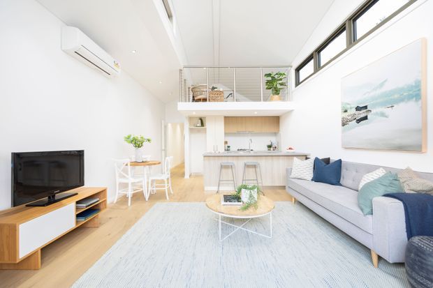 Bright open-plan living space with a contemporary kitchen and island, dining nook, and lounge area. The room features white walls and ceiling, pale timber flooring, a light grey sofa with blue and teal cushions, a pale rug, white dining furniture, timber TV cabinetry, high clerestory windows, and a mezzanine level with a metal balustrade. An abstract coastal-style artwork hangs above the sofa.