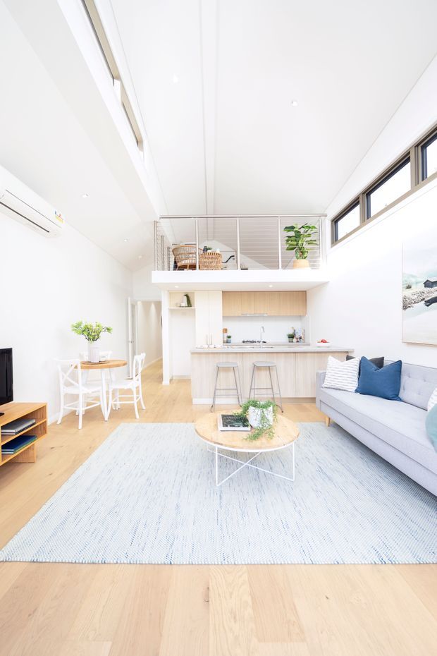 Bright, airy open-plan living room with a compact kitchen and dining area beneath a mezzanine. The space has white walls and ceiling, pale timber flooring, light kitchen cabinetry, a grey sofa with blue and white cushions, a pale textured rug, and a round coffee table. High-level windows and a simple metal mezzanine balustrade bring in abundant natural light.