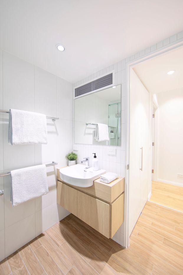 Bright, compact contemporary bathroom with white tiled walls, a timber-look tiled floor, floating light-wood vanity, white vessel basin, large mirrored cabinet, heated towel rails, and a glass shower enclosure reflected in the mirror. The bathroom opens to a light-wood-floored hallway through a white door.