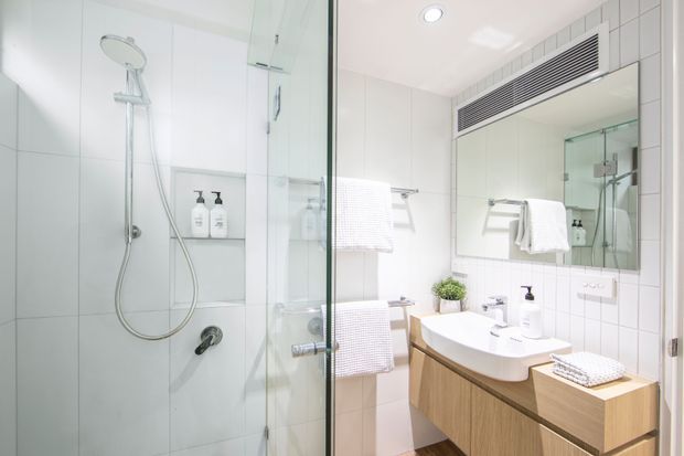 Bright contemporary bathroom with a glass-enclosed walk-in shower, white tiled walls, recessed shower niche, wall-mounted rain and handheld shower fittings, mirrored vanity, and floating timber-look cabinetry with a white integrated basin. A towel rail, folded towels, toiletries, and a small plant provide functional details.